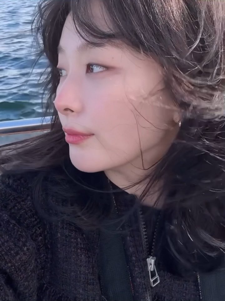 A close-up portrait of a young woman with long dark hair looking out over water, wearing a black zip-up jacket with the wind blowing through her hair.