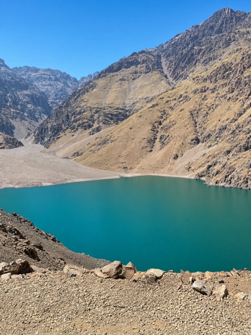 A scenic landscape photograph of a bright turquoise alpine lake surrounded by rocky, barren mountains under a clear blue sky, with a rocky shoreline in the foreground.