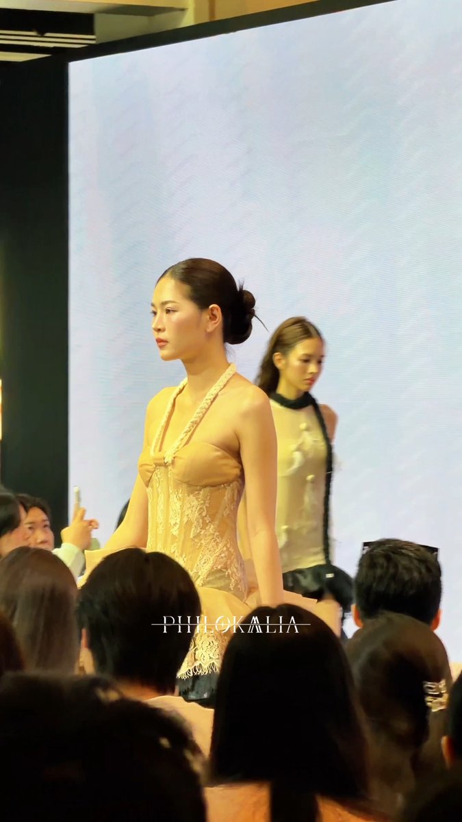 A fashion runway scene showing a model with her hair in a bun walking in a beige lace dress, with 'PHILOKALIA' branding visible and audience members in the foreground.