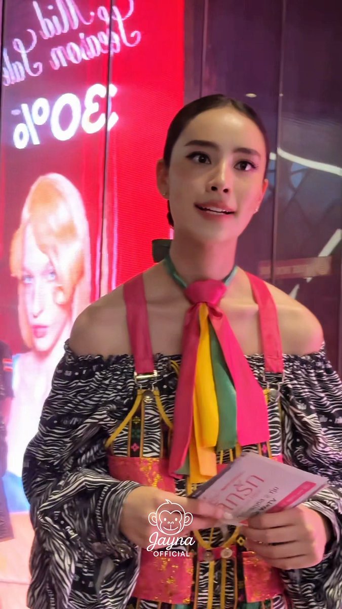 A woman in a colorful traditional-inspired outfit with a bright pink and green neck tie holds a booklet at an event, with a 'Hedi Slimane' / '30.04' sign reflected in the background.