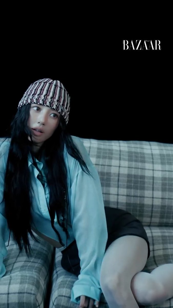 A moody, dimly lit editorial photo for Harper's Bazaar featuring a young woman with long dark hair wearing a striped beanie and an oversized blue shirt, posing on a plaid couch.