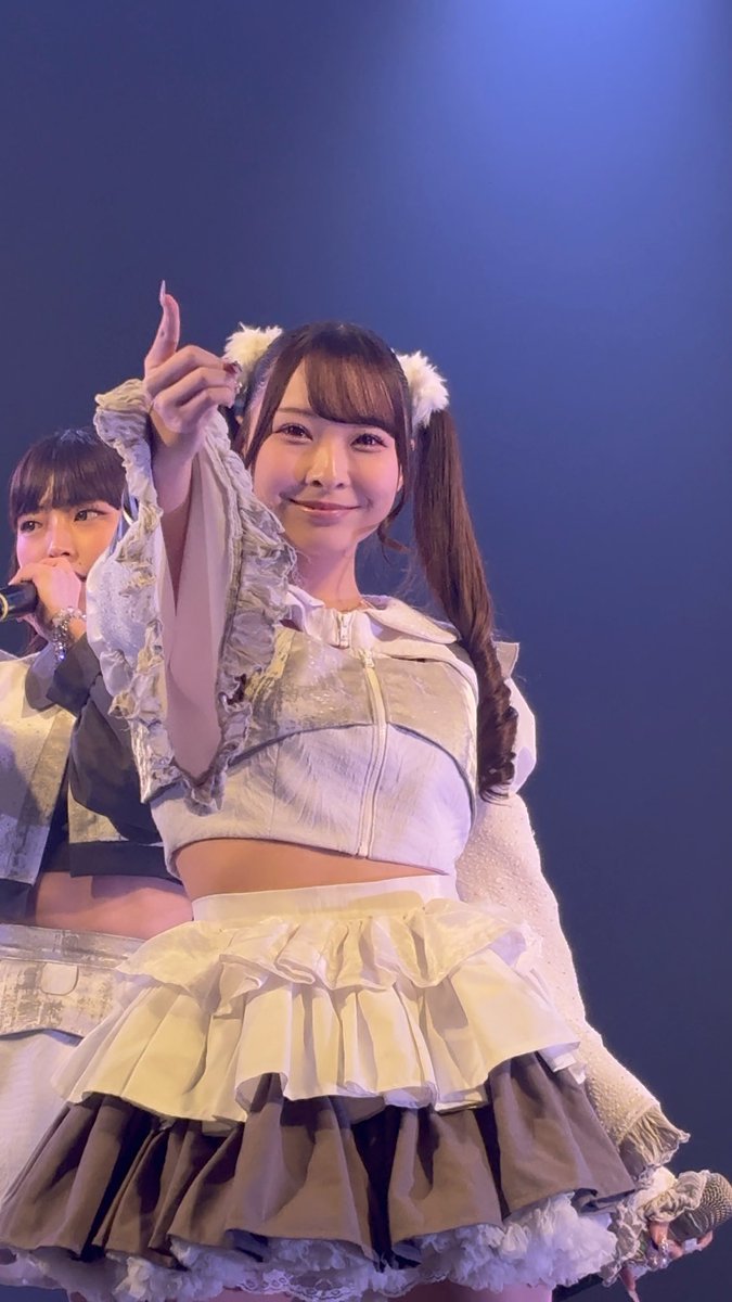 A young woman on stage in a silver crop top with white ruffled sleeves and a layered white and black skirt, wearing fluffy ear accessories and pointing upward with a smile.