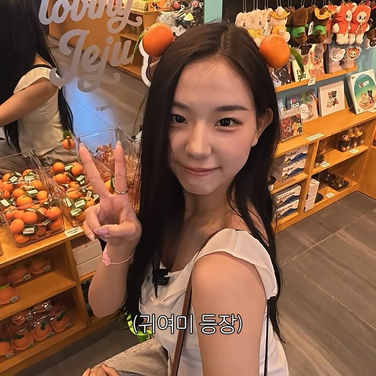 A young woman wearing an orange headband with fruit decorations takes a selfie in a shop, making a peace sign, with text overlay reading '(귀여미 등장)'.