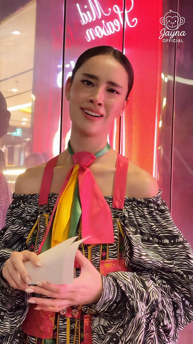 A woman smiles while holding what appears to be a red envelope, wearing a colorful scarf and patterned outfit, with a 'JAYNA OFFICIAL' monkey logo in the corner and a neon sign in pink/red behind her.