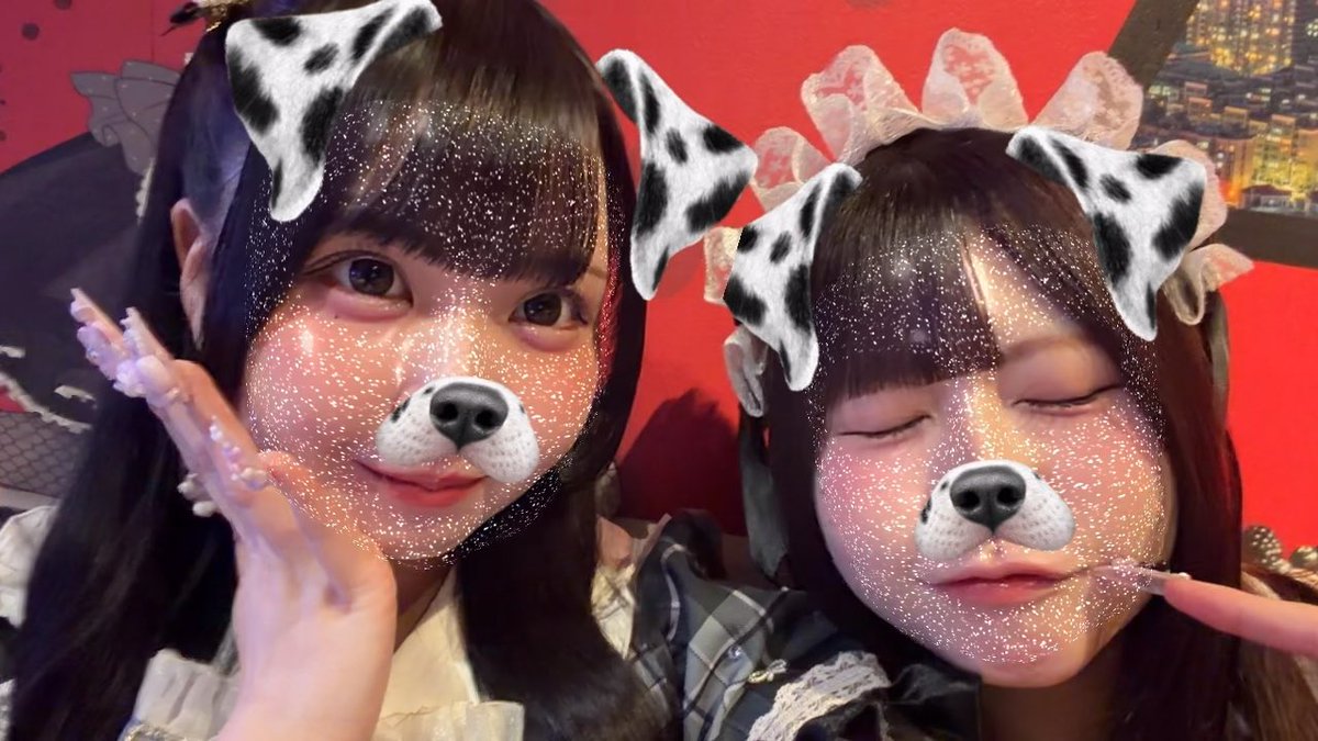 Selfie von zwei jungen Frauen mit einem Hundegesicht-Filter (sogenannter Dog-Face-Filter), die schimmerndes Make-up und gemusterte Schleifen tragen und vor einem roten Hintergrund posieren.