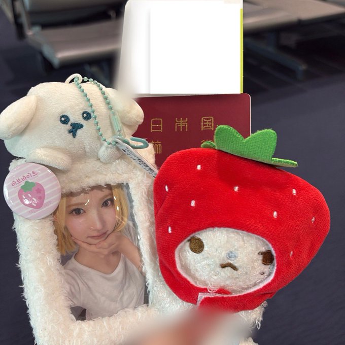 A phone case features a photo of a woman, topped with a white dog plushie, next to a red strawberry plush holding a passport with a Japanese flag design.