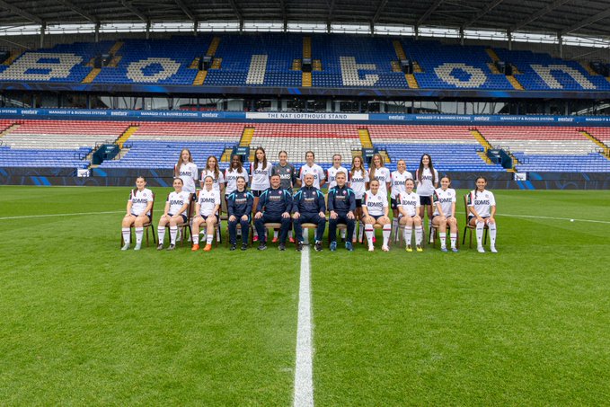 A sports team poses for a group photo on the field at a stadium with blue seating that spells out "BRISTOL" in the stands, with a banner reading "THE NAT LOFTHOUSE STAND".