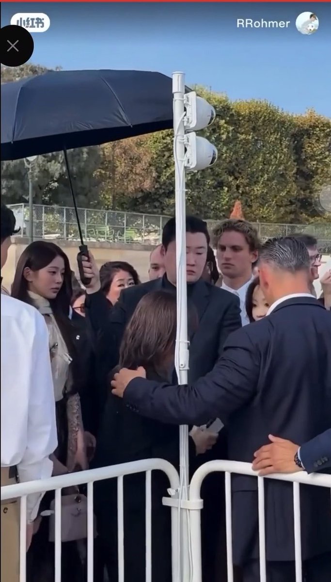 A crowd gathers behind a metal barricade, where a young woman in dark clothing appears to be interacting closely with several well-dressed individuals, including what looks like K-pop idols, while a person holds a black umbrella overhead to shield them—supporting the post's claim that she was in the front row directly engaging with the group rather than with lesser-known figures. The visible usernames "小红书" (Xiaohongshu) and "RRohmer" indicate the screenshot originates from a social media post.