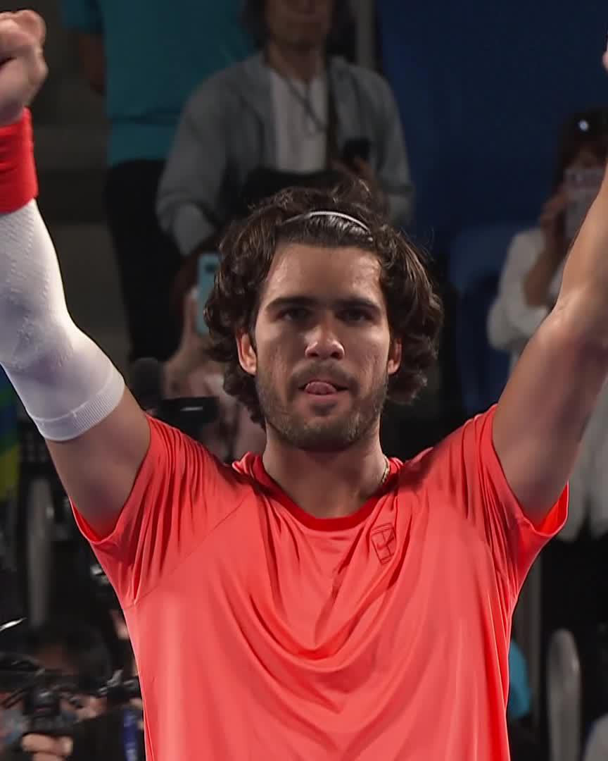 Tennis player Taylor Fritz, wearing a red shirt and a white wristband, raises his arms in celebration with his tongue out.
