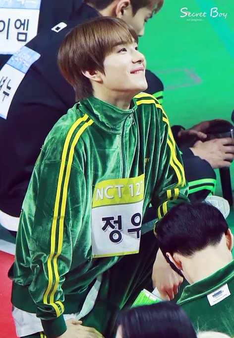 A young man in a green velour tracksuit featuring 'NCT 12' and Korean text '정우' (Jungwoo) smiles while participating in what appears to be an ISAC athletic event.
