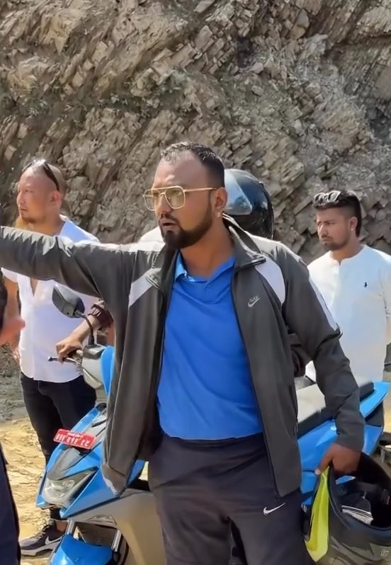 A bearded man wearing sunglasses, a blue polo shirt, and a gray Nike jacket pointing to the side while standing in front of a blue motorcycle, with other men and a rocky hillside in the background.