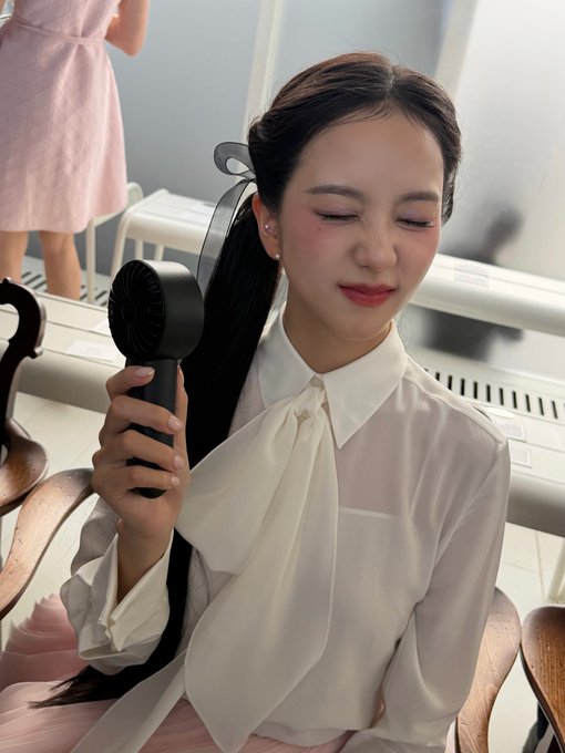 A young woman with a black ribbon in her hair smiles with her eyes closed while holding a small black handheld blower, dressed in a white blouse and pink skirt.