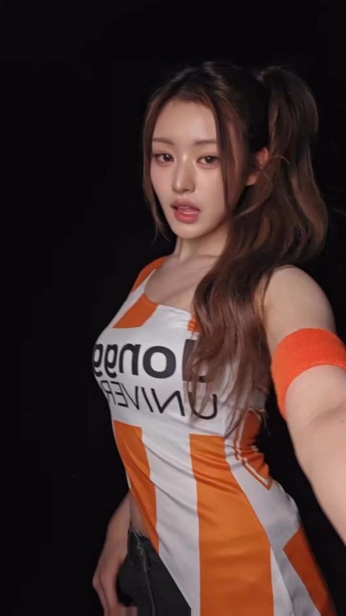 A woman with long wavy hair in pigtails poses for a selfie in a dark setting, wearing a tight orange and white striped crop top that reads 'londee' and an orange armband.