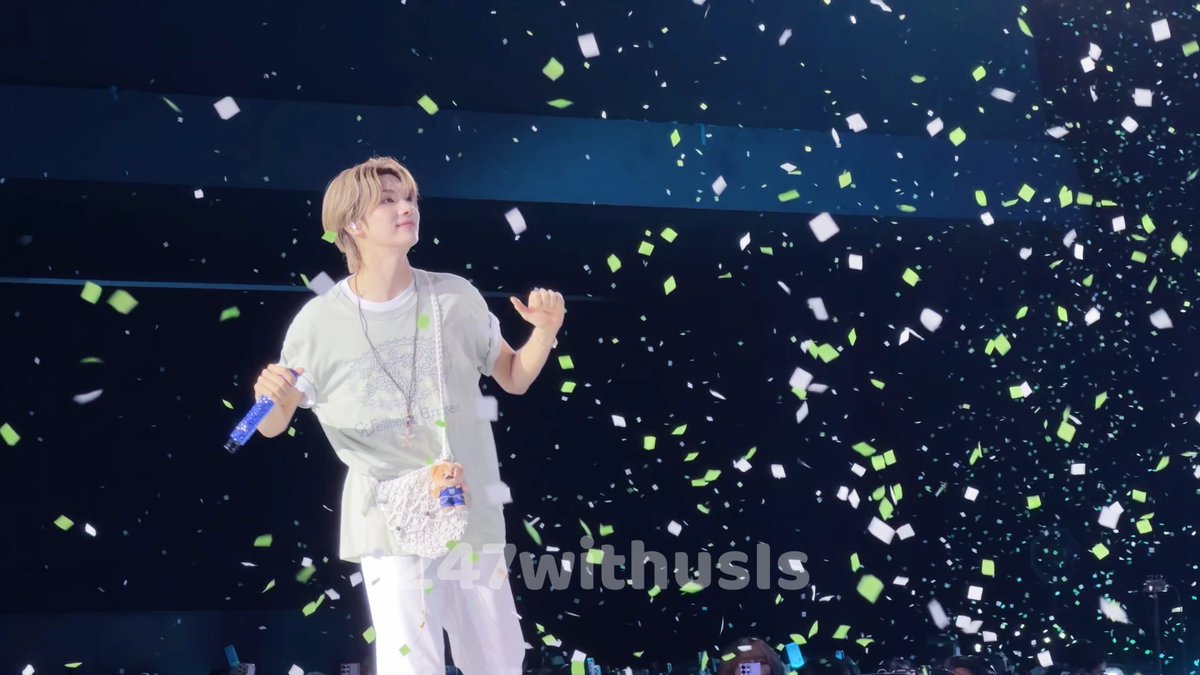 A performer on stage holding a microphone, surrounded by falling green and white confetti, with the text "247withusls" visible.