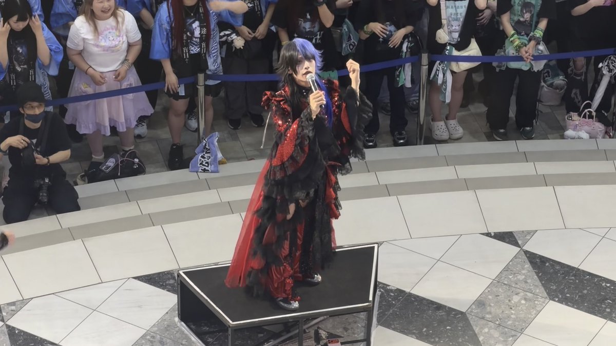 A performer in an elaborate red and black ruffled costume with a microphone, standing on a small stage in front of a crowd of onlookers.