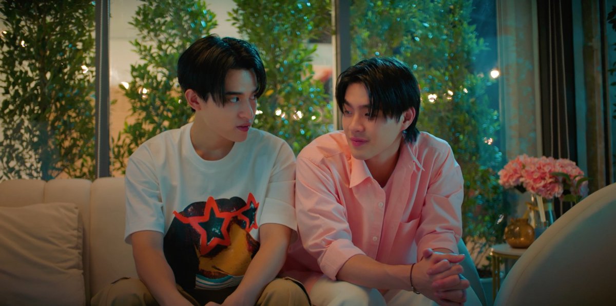 Two young men sit closely together on a beige couch in a warmly lit room with greenery behind them; one wears a graphic t-shirt with a red star design while the other wears a pink button-up shirt. The accompanying post suggests this is a moment from "YouManiacSeriesEP6" where the characters have just confessed their feelings and are considering starting a relationship, reflecting the intimate and emotionally charged atmosphere captured in the image.
