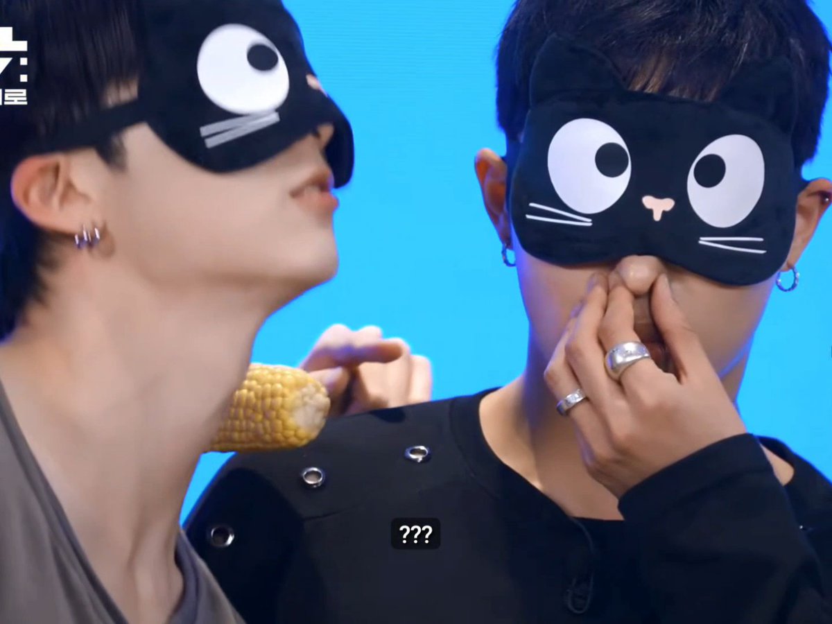 Two members of P1Harmony wear black cat eye masks while one playfully holds a corn cob up to another's face, humorously captioning the chaotic guessing moment with "???" displayed on screen.
