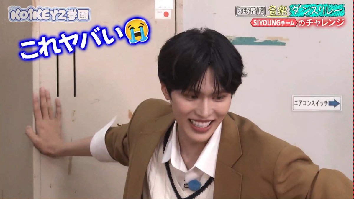 A young man with black hair, wearing a tan blazer over a white collared shirt and a v-neck sweater, is smiling excitedly while pressing his hand against a door frame, with Japanese text overlays reading "KO1KEYZ学園" and "これヤバい" alongside a crying-laughing emoji and "SIYOUNGチーム" graphics. His energetic, joyful expression and animated posture perfectly match the post's description of getting hyped by someone's cute side-to-side sway.