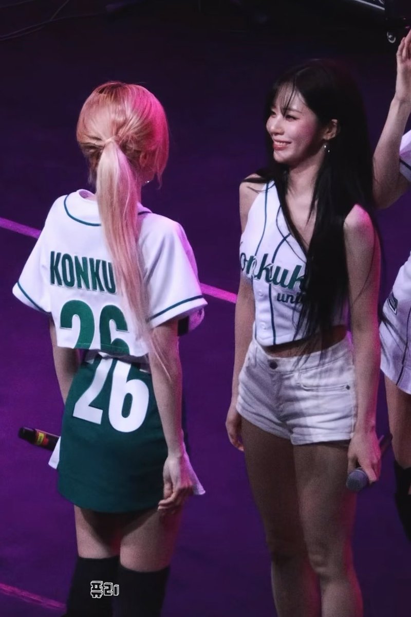 Two female K-pop idols on stage — one in a "KONKU 26" baseball jersey and skirt (back to camera), and another smiling at her in a matching "konku" cheerleader outfit holding a microphone.