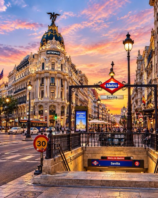 A vibrant sunset view of the Metropolis Building in Madrid, Spain, with the Sevilla Metro station entrance in the foreground.