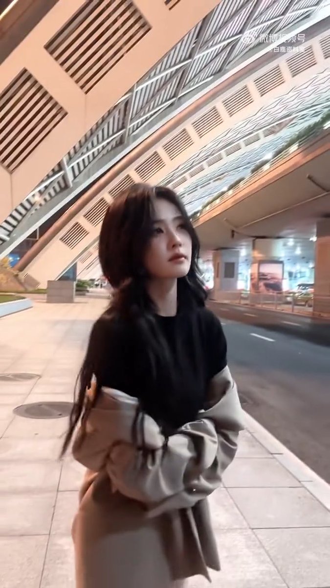 A young woman with long dark hair, wearing a black top and grey jacket, walks under a modern architectural overpass at dusk.