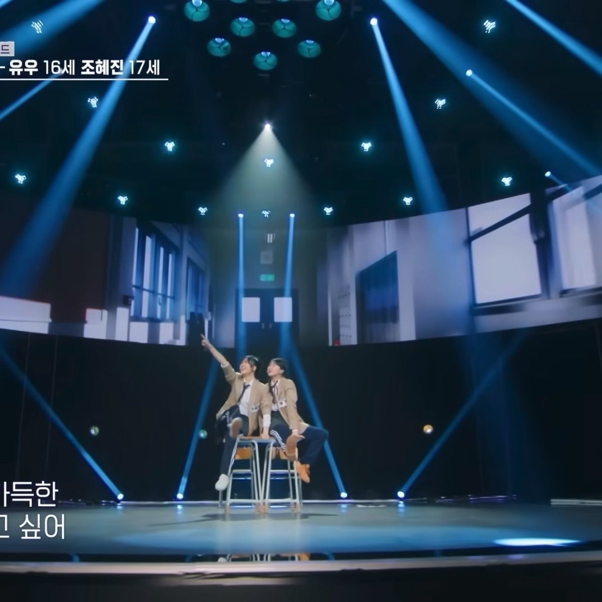 Two performers sit on stools on a brightly lit stage with blue spotlights, with Korean text visible reading '유우 16세 조혜진 17세' and partial subtitles '사특한' and '1 싶어'.