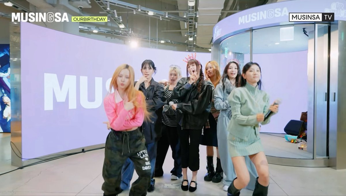 A group of seven young women in trendy outfits pose playfully for a MUSINSA TV broadcast, with 'MUSINGSA' and 'OURBIRTHDAY' text displayed on the large screen behind them.