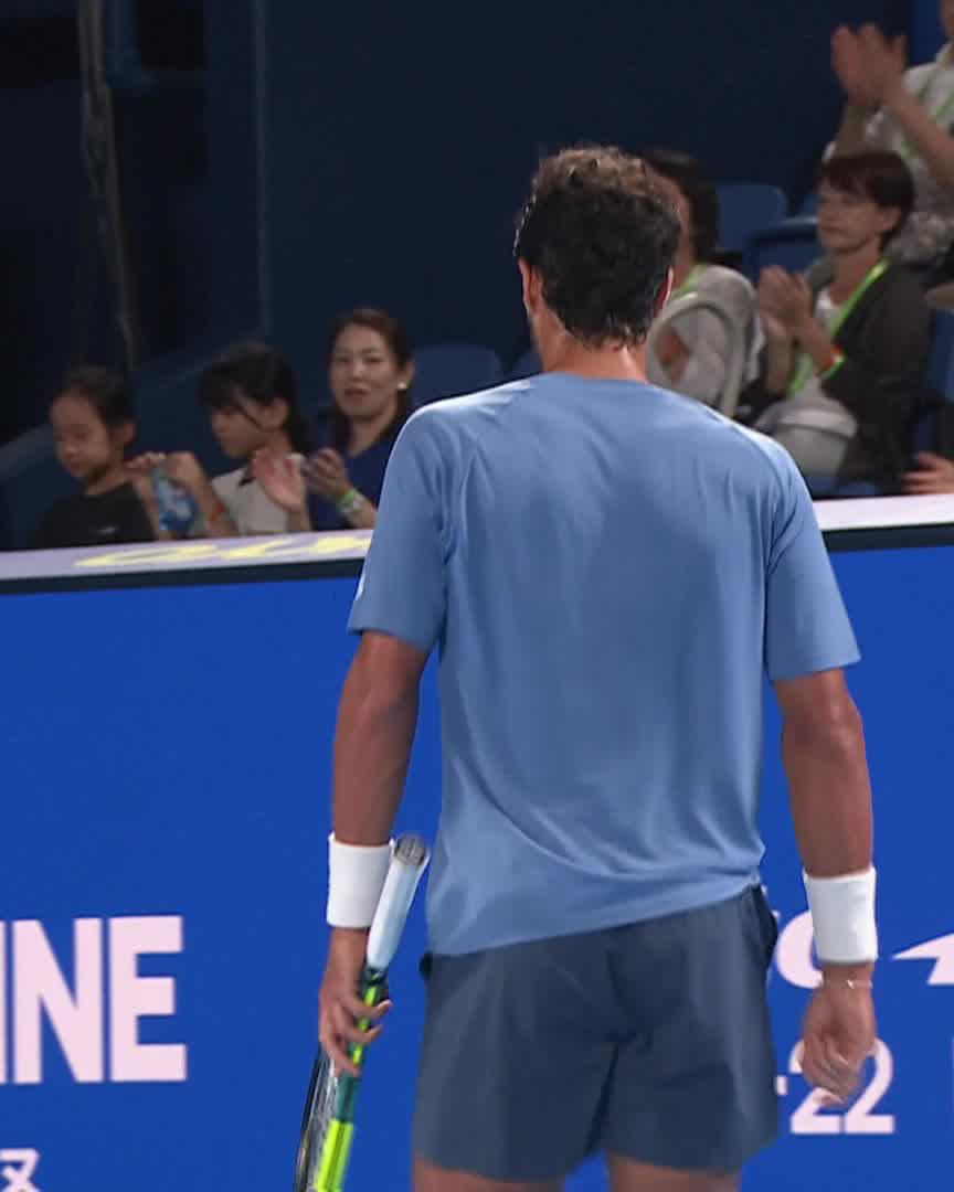 A male athlete in a light blue shirt and gray shorts stands holding a tennis racket, facing a crowd of clapping spectators at the Japan Open Tennis tournament, as indicated by the partial "INE" and "22" text on the blue backdrop. The scene captures the moment after Adolfo Daniel Vallejo's dominant first set victory over Jodar, with the enthusiastic crowd acknowledging his strong performance.
