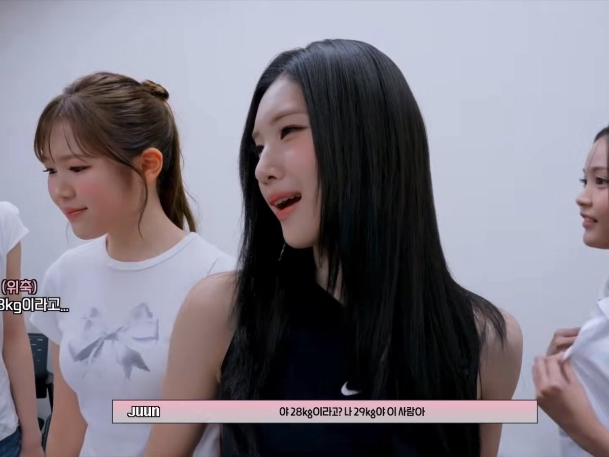 Three young women stand together in what appears to be a behind-the-scenes video; the center one wears a black Nike top and Korean subtitles read '아 28kg이라고? 나 29kg야 이 사람아' (Oh 28kg? I'm 29kg, this person).