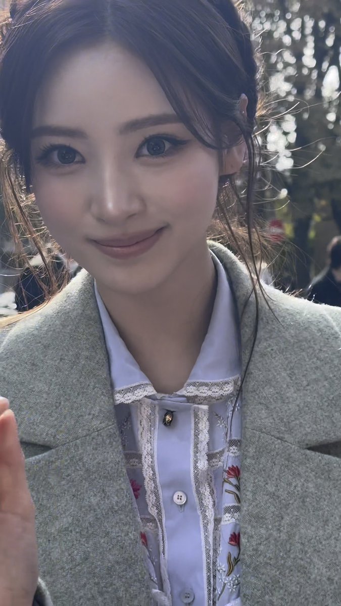 A young woman with dark hair and soft makeup smiles at the camera, wearing a white embroidered blouse with floral patterns under a light-colored blazer.