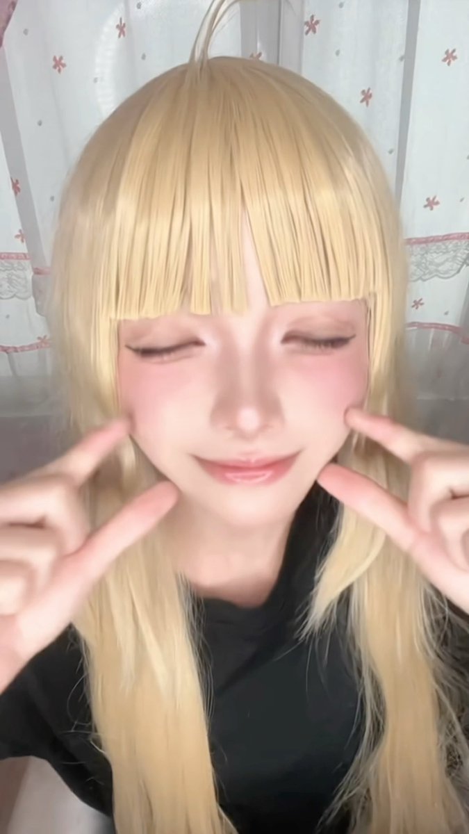 A person with long blonde hair and bangs poses with their eyes closed and fingers pressing on their cheeks.