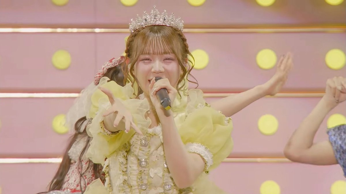 A female idol performer wearing a yellow princess-style gown and tiara sings into a microphone on stage, with another performer visible behind her.