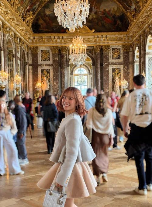 A smiling woman in a beige skirt suit stands in an ornate hall with chandeliers and painted ceilings, likely at a museum or palace, surrounded by other visitors.