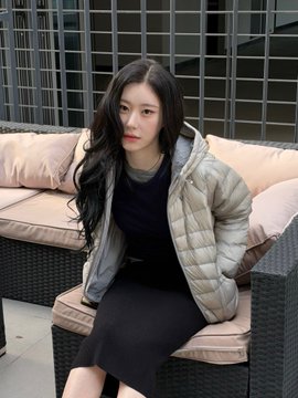 A young woman with long dark hair wearing a beige puffer jacket over a dark top sits on an outdoor wicker sofa.