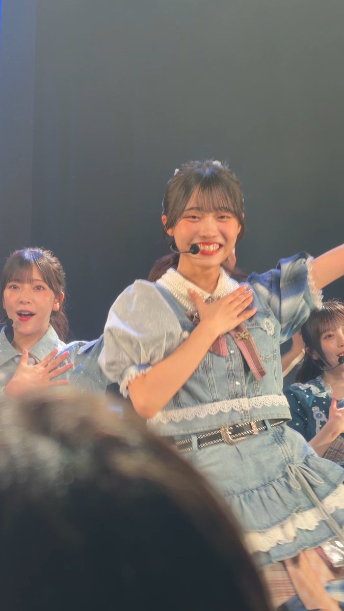 A young woman with dark hair smiling brightly on stage, wearing a denim and light blue outfit with a microphone headset, with other performers visible behind her.