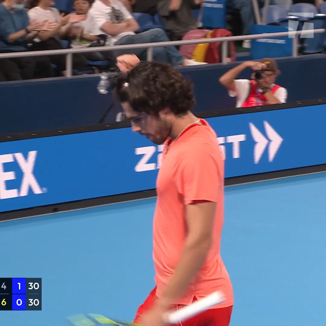 Carlos Alcaraz walks on the court during the Japan Open tennis tournament, with the scoreboard showing a tied score of 30-30, wearing his signature bright orange outfit as spectators watch from the stands.