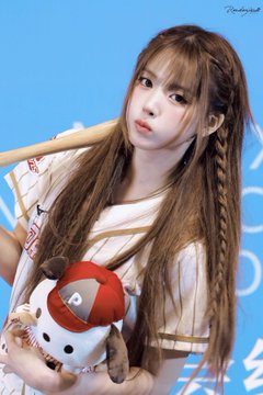 A young woman with long brown hair styled in braids holds a wooden baseball bat over her shoulder while cradling a small stuffed toy wearing a red baseball cap against a blue background.