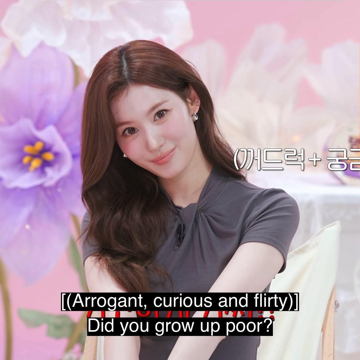 A woman with long brown hair wearing a grey short-sleeved top smiles during an interview, with on-screen text in Korean and English translating to 'Did you grow up poor?'.