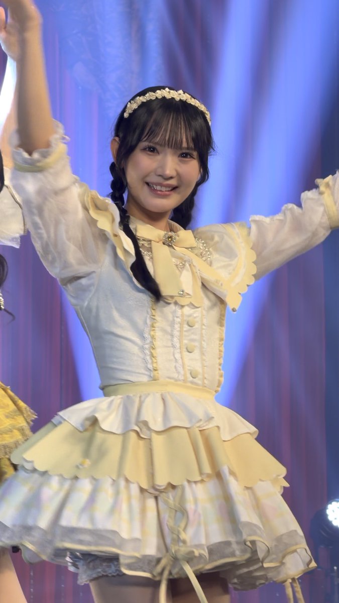 A young woman on stage in a yellow and white frilled idol dress with a pearl headband, smiling and waving with both arms raised.