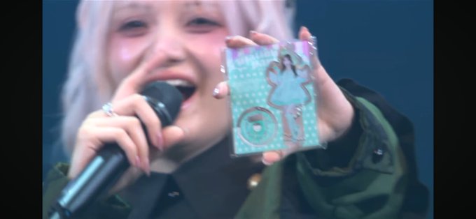 A performer with light-colored hair holds a microphone while displaying a decorative card or photo card featuring an illustrated figure in a blue dress. The "#宮野静" hashtag suggests this is from a stage or event appearance by Miyano Shizuka, likely a meet-and-greet moment where she shares merchandise with fans.