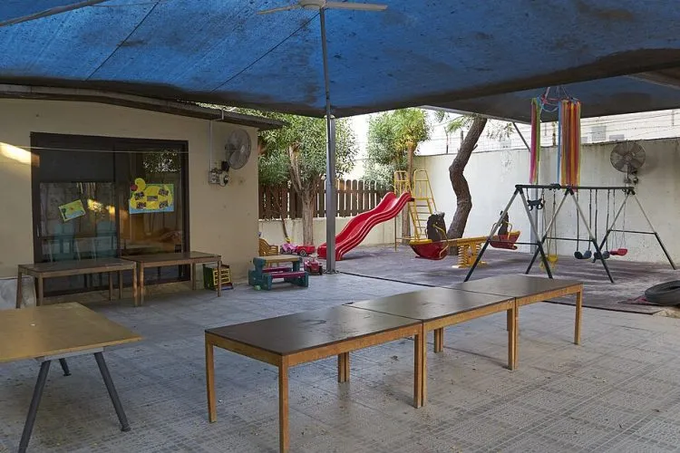 An outdoor covered play area with wooden benches, a red slide, swings, and a table, shaded by a large blue tarp.