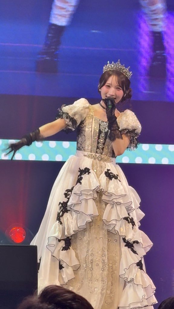 A performer on stage wearing an ornate white and black Victorian-style dress with a tiara and black gloves, holding a microphone during what appears to be an idol concert.