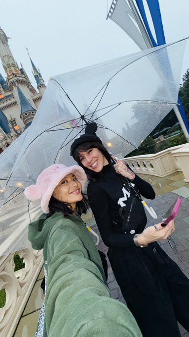 Two women take a selfie under a clear umbrella at what appears to be a Disney park, with Cinderella's Castle visible in the background.