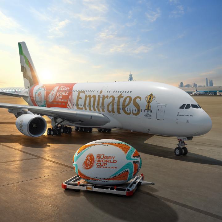 An Emirates Airbus A380, branded with "Rugby World Cup 2027 Australia" livery alongside a giant rugby ball on a cargo pallet, is displayed on the tarmac to mark the one-year countdown to the tournament. The aircraft's matching World Cup livery and the oversized ceremonial rugby ball reinforce Emirates' role as a sponsor ahead of the event in Australia.