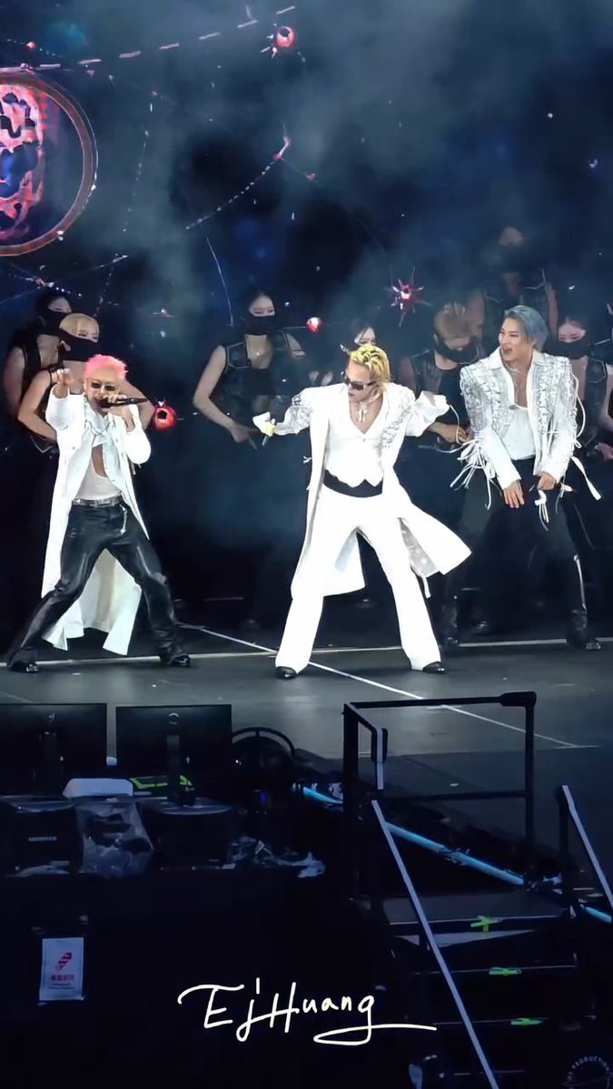 A concert stage performance by a K-pop group featuring performers in elaborate costumes (one with pink hair, one with blonde hair in white suit) with dancers in black behind them, watermarked 'EJ Huang'.