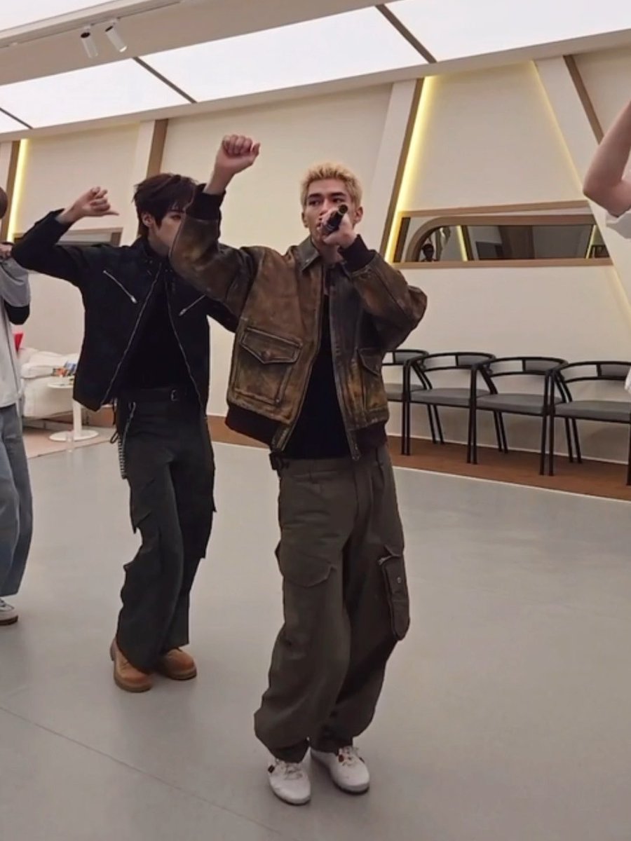 Two young men performing or dancing in an indoor space, one wearing a brown jacket and holding a microphone while the other poses with raised arms in a black jacket.