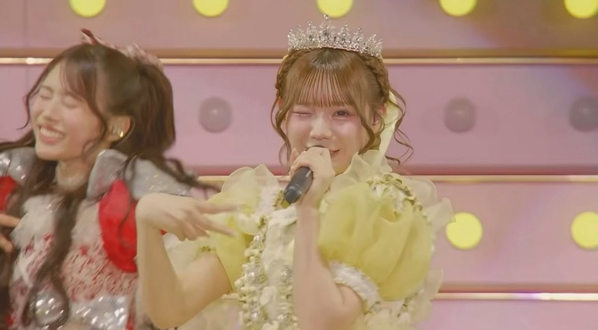 A J-pop or idol performance featuring a young woman in a yellow frilly dress and tiara singing into a microphone while winking, with another performer in red visible in the background.