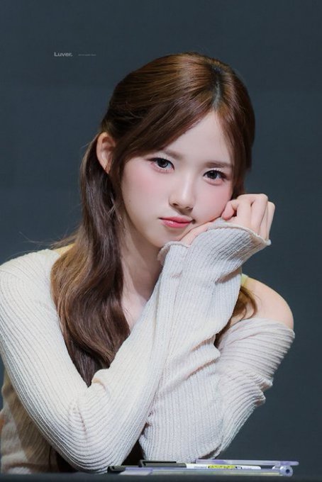 A close-up portrait of a woman with long brown hair wearing a cream-colored knit sweater, posing with her hand resting near her face against a dark gray background, watermarked 'Liver_'.