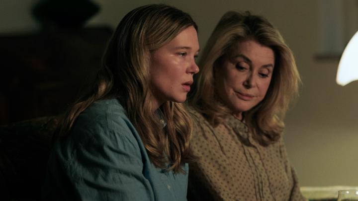 Two women with long blonde hair, one in a blue denim shirt and the other in a patterned blouse, sit close together with somber expressions in a dimly lit room, evoking the intimate, tense atmosphere of the thriller. The image captures a moment of shared concern between them, fitting the film's theme of trust and deception.
