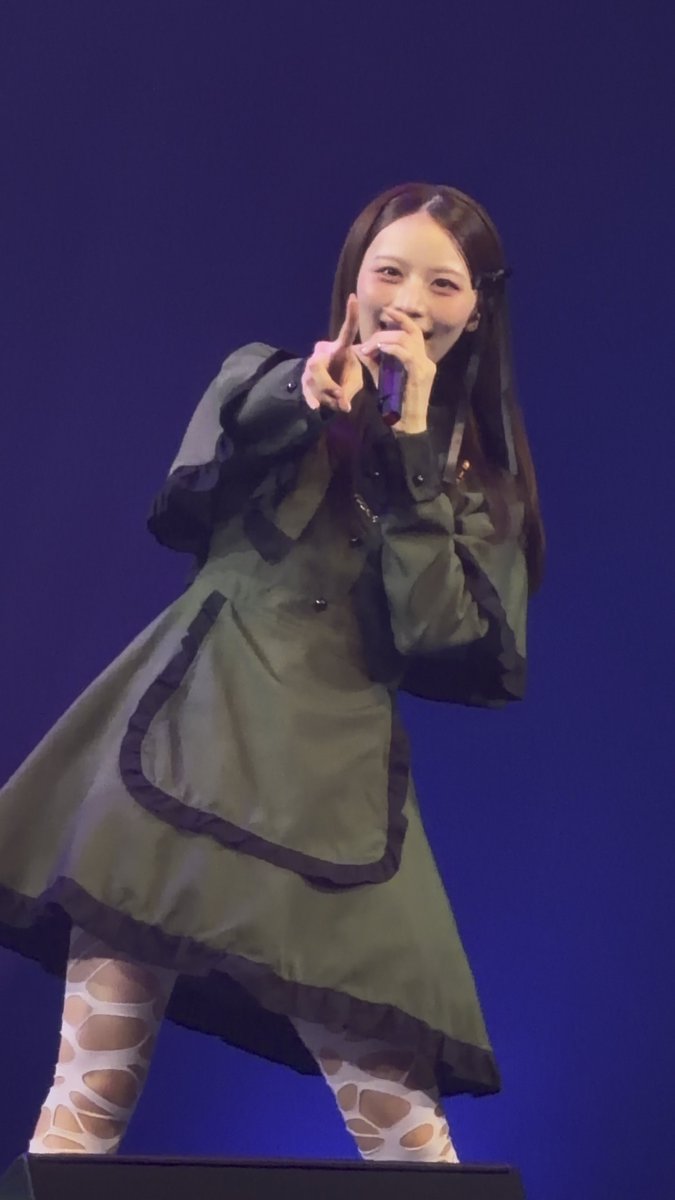 A female performer on stage against a blue background, singing into a microphone while wearing an olive green ruffled dress and patterned stockings.